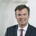 Mann mit dunklem Haar und Lächeln im Gesicht trägt ein schwarzes Sakko, ein weißes Hemd und eine dunkelrote Krawatte.