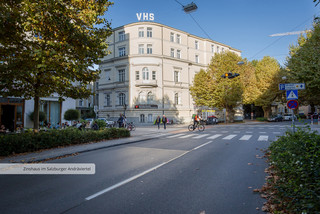 Straßenkreuzung in der Stadt Salzburg mit grünen Bäumen, Zebrastreifen, mehreren Passanten und einem Radfahrer. Ein großes historisches Gebäude mit weißer Fassade und 5 Stockwerken, welches am Dach die Aufschrift "VHS" trägt.
