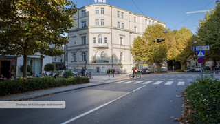 Straßenkreuzung in der Stadt Salzburg mit grünen Bäumen, Zebrastreifen, mehreren Passanten und einem Radfahrer. Ein großes historisches Gebäude mit weißer Fassade und 5 Stockwerken, welches am Dach die Aufschrift "VHS" trägt.