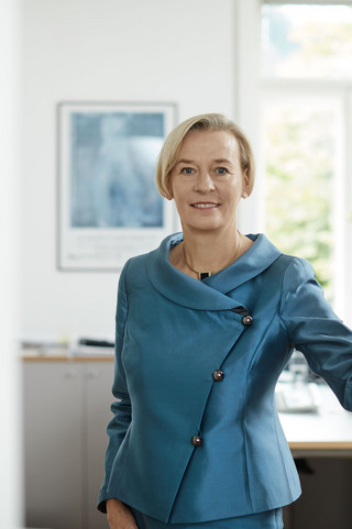 Eine Beraterin trägt ein hellblaues Kostüm und lächelt. Sie befindet sich in einem hellen Büro des Bankhaus Spängler.