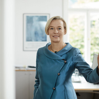 Eine Beraterin trägt ein hellblaues Kostüm und lächelt. Sie befindet sich in einem hellen Büro des Bankhaus Spängler.