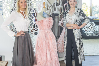 Zwei lachende Frauen stehen auf einem Podest. In der Mitte befindet sich ein Designerkleid mit Federn in rosa Farbe. Eine Frau hat blonde lange Haare, sie trägt eine weiße Bluse und einen grauen Plisseerock. Die andere Frau trägt eine schwarze Hose, ein weißes Top und einen lange weiß-schwarze leichten Bluse. Sie hat schwarze Haare und trägt sie zusammengebunden.