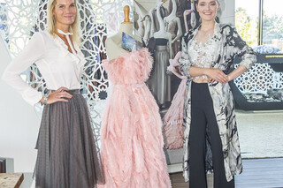 Zwei lachende Frauen stehen auf einem Podest. In der Mitte befindet sich ein Designerkleid mit Federn in rosa Farbe. Eine Frau hat blonde lange Haare, sie trägt eine weiße Bluse und einen grauen Plisseerock. Die andere Frau trägt eine schwarze Hose, ein weißes Top und einen lange weiß-schwarze leichten Bluse. Sie hat schwarze Haare und trägt sie zusammengebunden.