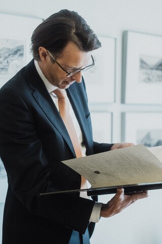 Ein Herr in Geschäftskleidung blättert vor einer mit Gemälden bestückten Wand in einem historischen Buch.