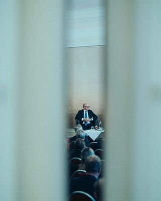 Blick durch einen Schlitz auf eine Bühne mit einer Podiumsdiskussion und deren Publikum.