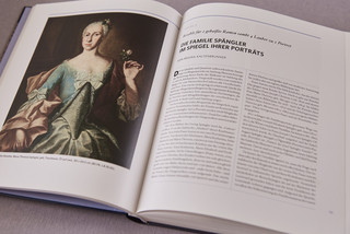 Aufgeklapptes Buch in A4-Format. Linksseitig ist ein Gemäldeportrait einer Dame mit hochgesteckten Haaren und opulentem Kleid zu sehen. Rechts befindet sich die Textseite mit der Überschrift "Die Familie Spängler im Spiegel ihrer Porträts".