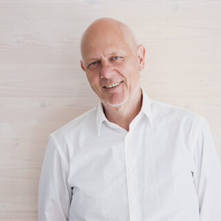Mann mit Glatze in weißem Hemd lächelt freundlich. Er steht vor einer weißen Holzwand, man sieht nur seinen Oberkörper.