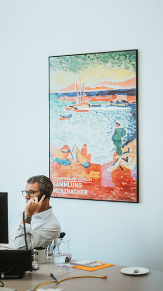 Ein telefonierender Herr sitzt an einem Schreibtisch vor einem Gemälde.
