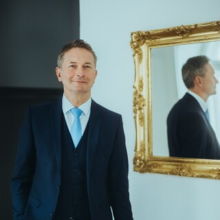 Ein Mann im Anzug und mit Krawatte steht neben einem Spiegel mit barockem, goldenem Rahmen.