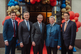 Fünf Herren in Geschäftskleidung stehen vor einem mit Luftballonen geschmückten Portal.