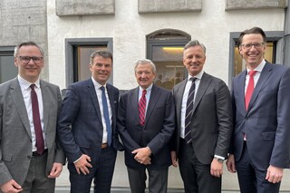 Fünf Herren in Geschäftskleidung stehen lächelnd vor einer Hausfassade.