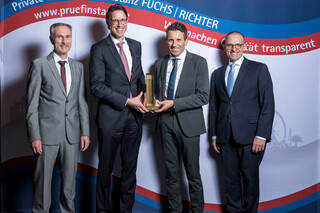 Vier lächelnde Herren in Anzug posieren mit einer Trophäe vor einer Fotowand.