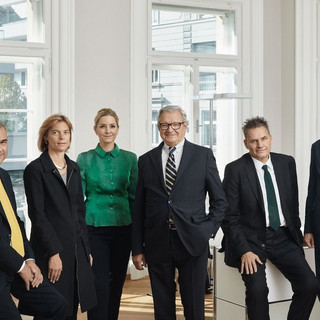 6 Personen (2 Frauen, 4 Männer) in einem hellen Büro. 5 Personen tragen dunkle Kleidung, 1 Frau trägt ein dunkelgrünes Oberteil.
