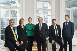 6 Personen (2 Frauen, 4 Männer) in einem hellen Büro. 5 Personen tragen dunkle Kleidung, 1 Frau trägt ein dunkelgrünes Oberteil.