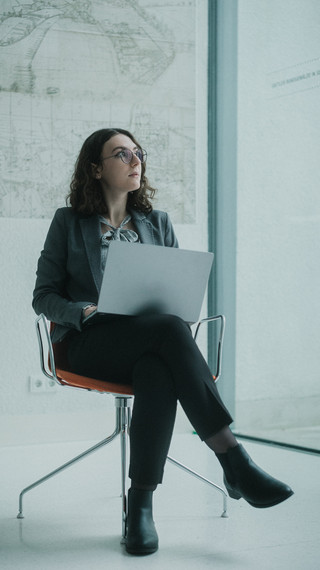 Eine Frau mit Brille sitzt auf einem Stuhl, auf ihrem Schoß liegt ein Laptop. 