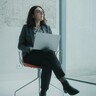 Eine Frau mit Brille sitzt auf einem Stuhl, auf ihrem Schoß liegt ein Laptop. 