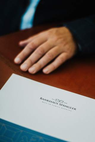 Eine Broschüre mit der Aufschrift "Bankhaus Spängler" liegt neben einer Hand auf einem Tisch.