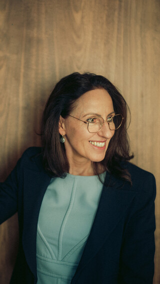 Eine Frau mittleren Alters mit dunklen schulterlangen Haaren, hellblauem Oberteil und dunklem Blazer blickt nach rechts; links im Bild steht eine Blumenvase.