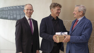 Drei Männer vor einer Wand mit der Aufschrift "Bankhaus Spängler". Ein Mann überreicht dem anderen ein Geschenk.