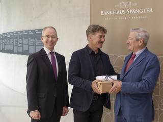 Drei Männer vor einer Wand mit der Aufschrift "Bankhaus Spängler". Ein Mann überreicht dem anderen ein Geschenk.
