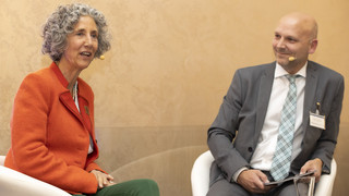 Eine Frau und ein Mann diskutieren am Podium, den Hintergrund ziert eine beige-gemusterte Wand. Die Frau hat gräuliche Locken und trägt einen orange-farbigen Blazer und eine grüne Hose. Der Mann trägt einen grauen Anzug, ein weißes Hemd und eine blau-grau karierte Krawatte.