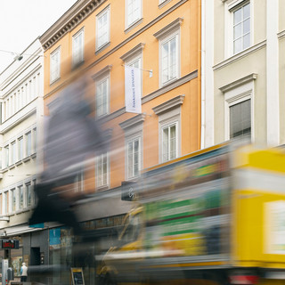 Geschäftiges Treiben der der Niederlassung Innsbruck des Bankhaus Spängler. Die Fassade ist orange, das Bild allgemein verzerrt.