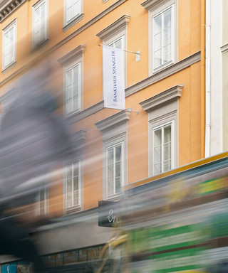 Geschäftiges Treiben der der Niederlassung Innsbruck des Bankhaus Spängler. Die Fassade ist orange, das Bild allgemein verzerrt.