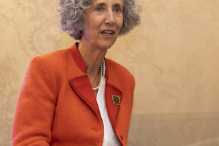 Frau mit grauen Locken sitzt auf einem beigen Stuhl. Sie spricht gerade und trägt einen orange-farbigen Blazer, eine weiße Bluse und eine dunkelgrüne Hose. Den Hintergrund ziert eine beige-gemusterte Wand.