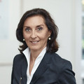 Frau mit welligen Haaren lächelt freundlich. Sie trägt einen dunklen Blazer und eine weiße Bluse sowie weiße Ohrringe. Ihre Brille hält sie in der Hand.