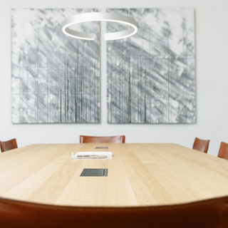 Besprechungsraum mit einem hellen, eckigen Holztisch. Mehrere dunkelbraune Stühle sind darum platziert. Eine graue, rundliche Leuchte hängt von der Dicke über die Mitte des Tisches. Zwei grau-weiße Bilder mit Strichen hängen an der Wand.