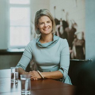 Eine Dame in einem Kleid sitzt lächelnd an einem Tisch vor einem Rollup.