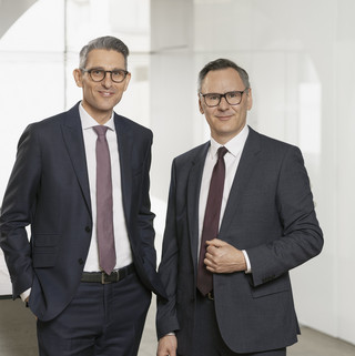 Rudolf Eder (r.) und Martin Kaufmann Zwei Herren im Anzug und Krawatte mit Brille stehen vor einer Glaswand in einem modernen Gebäude
