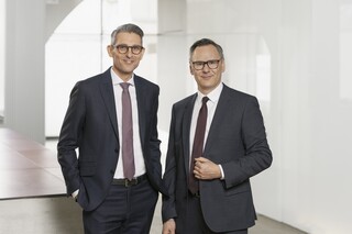 Zwei Herren im Anzug und Krawatte mit Brille stehen vor einer Glaswand in einem modernen Gebäude