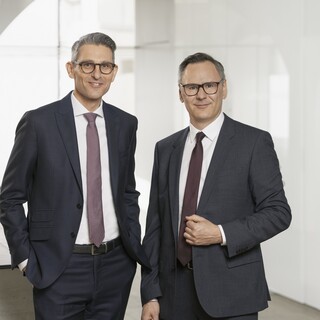 Rudolf Eder (r.) und Martin Kaufmann Zwei Herren im Anzug und Krawatte mit Brille stehen vor einer Glaswand in einem modernen Gebäude