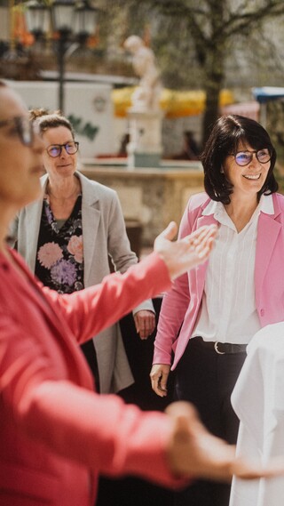 Eine Dame gestikuliert mit den Händen vor zwei lächelnden Damen und einem Brunnen.