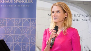 Frau mit blonden Haaren und pinkem Kleid begrüßt die Gäste und hält ein Mikrofon in der Hand.
