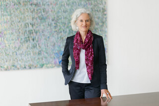 Lächelnde Frau mit braunem, lockigem Haar steht bei Türeingang zu einem Büro. Sie trägt eine weißes Bluse, einen dunkelrosa Blazer und eine schwarze Hose sowie eine weiße Kette. Im Hintergrund sieht man einen Stuhl und einen runden Tisch.