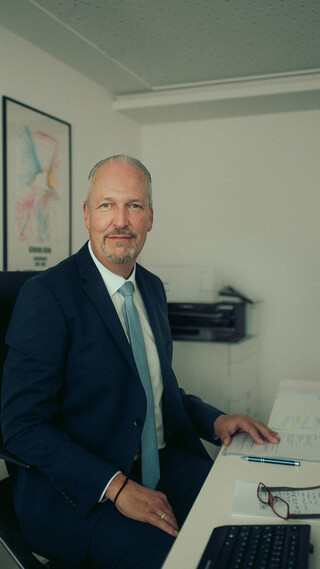 Ein Mann mit grauem Bart und Haar, blauer Krawatte und dunkelblauem Anzug sitzt an seinem Schreibtisch und blickt in die Kamera.