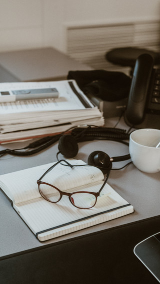 Auf einem Tisch liegt eine Brille auf einem Heft, daneben Kopfhörer und eine Kaffeetasse.
