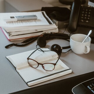 Auf einem Tisch liegt eine Brille auf einem Heft, daneben Kopfhörer und eine Kaffeetasse.