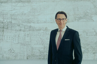 Mann mit Brille steht in einem Büro und lehnt sich an ein Pult an. Er trägt einen dunklen Anzug, ein weißes Hemd und eine hellrote Krawatte. 