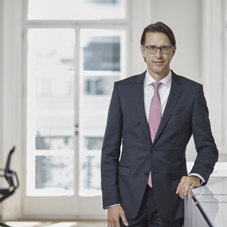 Mann mit Brille steht in einem Büro und lehnt sich an ein Pult an. Er trägt einen dunklen Anzug, ein weißes Hemd und eine hellrote Krawatte.