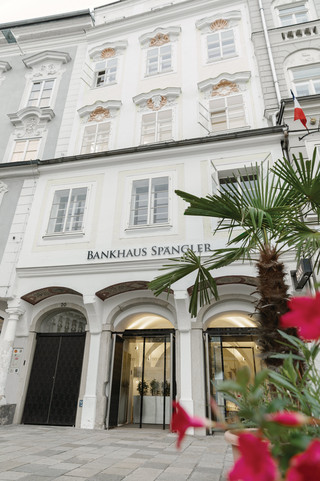 Bankgebäude der Niederlassung in Linz des Bankhaus Spängler von außen. Helles Gebäude mit 4 Stockwerken, im Eingangsbereich sind Glastüren. Ebenfalls im Bild ersichtlich ist eine Palme und rote Blumen.
