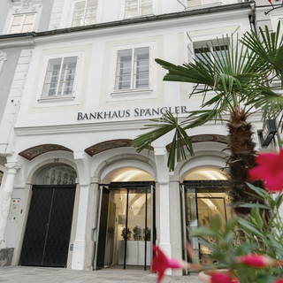 Bankgebäude der Niederlassung in Linz des Bankhaus Spängler von außen. Helles Gebäude mit 4 Stockwerken, im Eingangsbereich sind Glastüren. Ebenfalls im Bild ersichtlich ist eine Palme und rote Blumen.