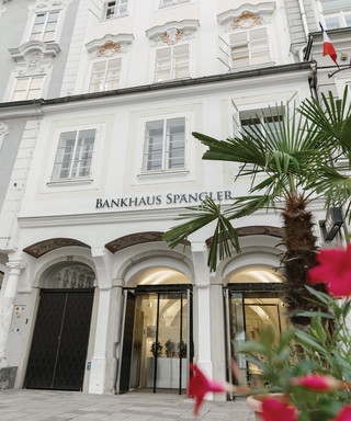 Bankgebäude der Niederlassung in Linz des Bankhaus Spängler von außen. Helles Gebäude mit 4 Stockwerken, im Eingangsbereich sind Glastüren. Ebenfalls im Bild ersichtlich ist eine Palme und rote Blumen.