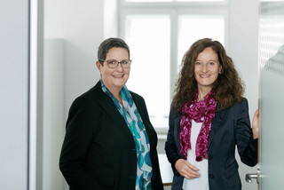 2 Frauen stehen in einem Büro neben einer Glastür, sie lächeln freundlich und tragen schwarze Blazer. Die Dame links hat kurze Haare, trägt eine Brille und ein blaues Tuch. Die Dame rechts hat lange lockige Haare und trägt ein dunkelrotes Tuch.