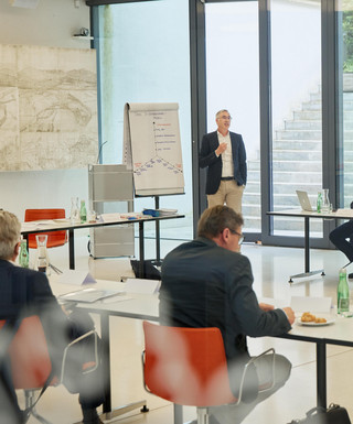 Personen in Geschäftskleidung sitzen in U-Form und lauschen einem Vortragenden mit Flipchart.