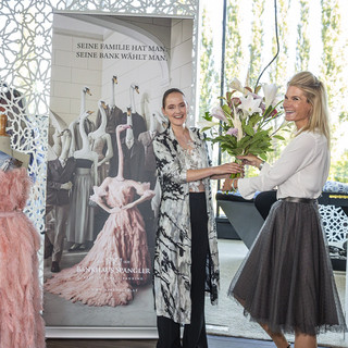 Zwei lachende Frauen, die gemeinsam einen großen hellen Blumenstrauß in den Händen halten. Nebenbei findet sich ein Designerkleid mit Federn in rosa Farbe.