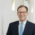 Mann mit braunen Haaren und rundlicher Brille lächelt höflich. Er öffnet gerade eine Tür und hält einen Kugelschreiber in der anderen Hand. Gekleidet ist er in ein weißes Hemd, ein schwarzes Sakko und eine hellblaue Krawatte.