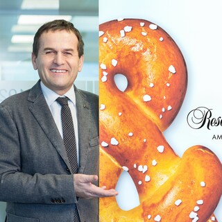 Zweigeteiltes Bild. Links ein Mann in grauem Anzug mit grauer Krawatte, der lächelt. Er hat braune Haare. Er winkelt seinen Ellbogen ab und deutet auf das andere Bild. Dort ist das Logo von "Resch & Frisch", mit einer Laugenbreze abgebildet.